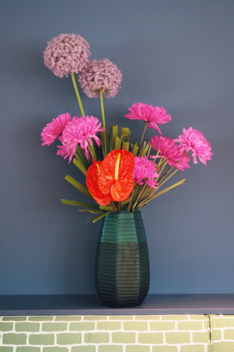 Grüne Blumenvase, welche auf der Rückwand einer Sitzbank steht mit violetten Kugelbumen, rosa Blumen und roten Anthurien