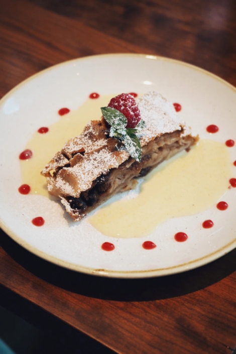 Nahaufnahme eines servierten Apfelstrudels mit Puderzucker, Vanillesauce und einer frischen Himbeere auf einem dekorativen Teller im Fräulein Wagner Restaurant in München.