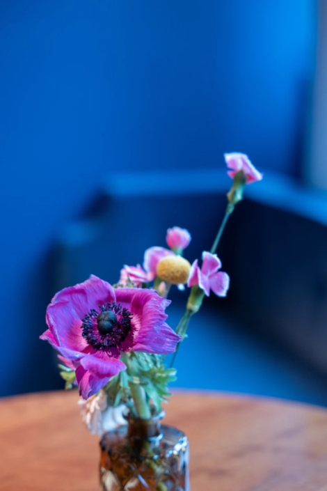 Vase mit zarten, pinken und gelben Blüten auf einem Holztisch, vor einem blauen Hintergrund im Fräulein Wagner Restaurant in München.