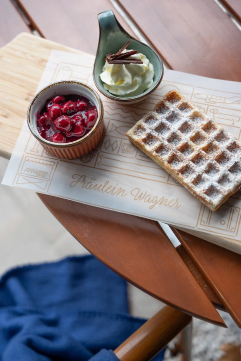 Hausgemachte Waffel mit Puderzucker, serviert mit Sahne und frischen Kirschen auf einer dekorativen Servierplatte im Fräulein Wagner Restaurant in München.